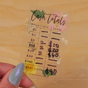 May include: A clear plastic cash register tracker with floral accents and gold foil. The tracker has a column for bills, quantity, and total. The totals are handwritten in black ink. The total cash is $90.