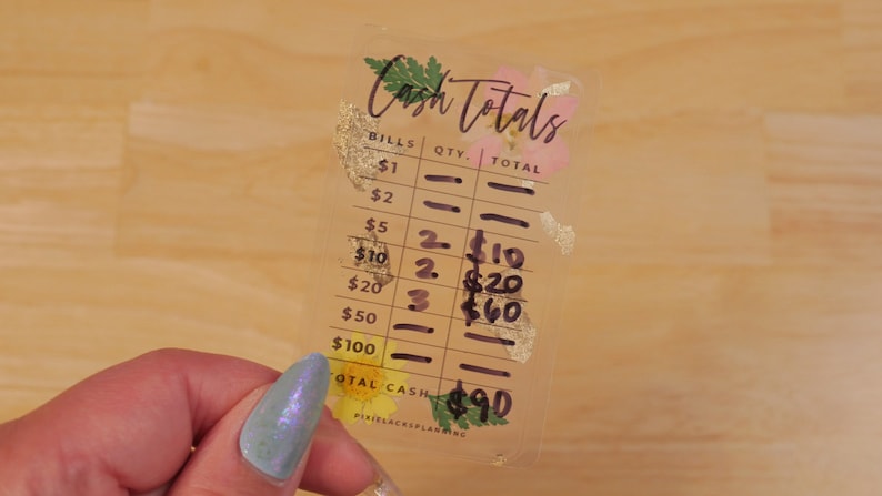 May include: A clear plastic cash register tracker with floral accents and gold foil. The tracker has a column for bills, quantity, and total. The totals are handwritten in black ink. The total cash is $90.