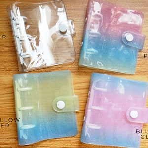 以下が含まれることがあります： 異なる色のグラデーションとグリッターが付いた透明なプラスチックバインダーが4つあります。バインダーには、「WHITE NO GLITTER」、「PINK BLUE YELLOW」、「BLUE YELLOW GLITTER」、「BLUE PINK GLITTER」とラベルが付けられています。