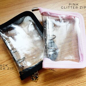 以下が含まれることがあります： 黒とピンクのグリッタージッパーが付いた透明なプラスチック製のジッパー付きポーチ2つ。ポーチは開いており、内部のバインダーリングシステムが見えます。