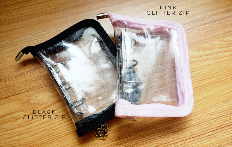 以下が含まれることがあります： 黒とピンクのグリッタージッパーが付いた透明なプラスチック製のジッパー付きポーチ2つ。ポーチは開いており、内部のバインダーリングシステムが見えます。