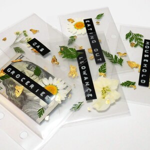 以下が含まれることがあります： 黒いラベルと金のアクセントが付いた透明なプラスチック製の封筒が5つ。ラベルには「ガソリン」、「食料品」、「手当」、「外食」、「家事」と書いてあります。各封筒には押し花と金箔が入っています。