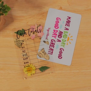 May include: A clear plastic cash register with a floral design and the text "Cash Totals" on the top. The register has columns for quantity and total. A white card with the text "Make a bad day good and a good day great!" is on top of the register.