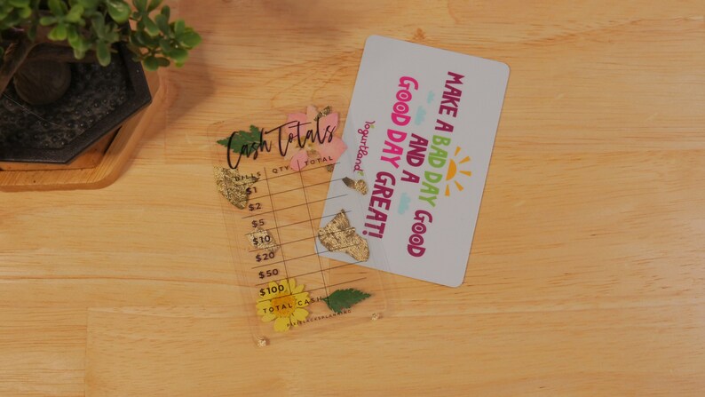 May include: A clear plastic cash register with a floral design and the text "Cash Totals" on the top. The register has columns for quantity and total. A white card with the text "Make a bad day good and a good day great!" is on top of the register.