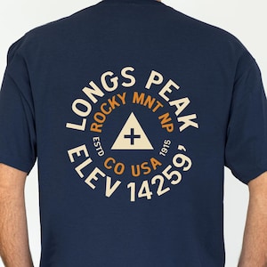 May include: A navy blue short-sleeve t-shirt featuring a circular graphic on the back. The text reads LONGS PEAK, ROCKY MNT NP, ESTD 1915, CO USA, and ELEV 14259'. A central triangle contains a plus sign. The design uses cream and orange lettering.