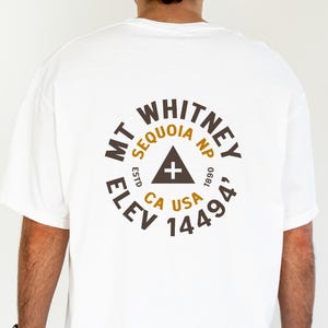 May include: White t-shirt with a circular graphic on the back featuring "MT WHITNEY SEQUOIA NP CA USA 14494" in brown text. The design includes a triangle and cross symbol. The shirt is worn by a person with black pants and a black watch.