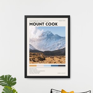 May include: A framed print of Mount Cook, New Zealand, with the text "CANTERBURY, NEW ZEALAND MOUNT COOK." The print features a mountain landscape with a color palette and is displayed above a black metal chair with yellow pillows and a potted plant.