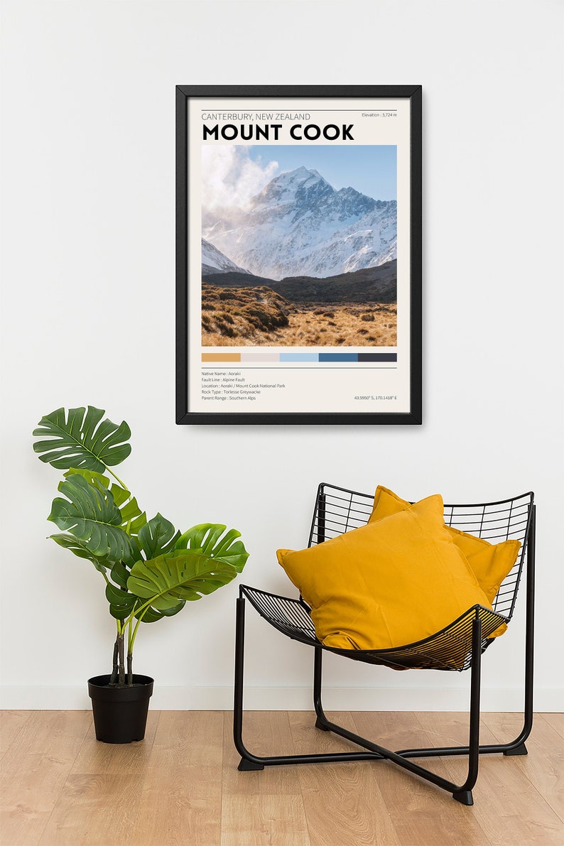 May include: A framed print of Mount Cook, New Zealand, with the text "CANTERBURY, NEW ZEALAND MOUNT COOK." The print features a mountain landscape with a color palette and is displayed above a black metal chair with yellow pillows and a potted plant.