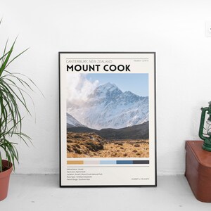 May include: A framed poster featuring Mount Cook in New Zealand, with the text "Mount Cook" in bold black letters. The image shows a snow-capped mountain and a field of brown grass. A green lantern and a brown suitcase are next to the poster.