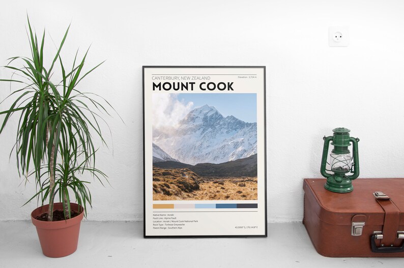 May include: A framed poster featuring Mount Cook in New Zealand, with the text "Mount Cook" in bold black letters. The image shows a snow-capped mountain and a field of brown grass. A green lantern and a brown suitcase are next to the poster.