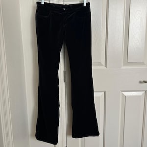 May include: Black velvet pants hanging on a hanger. The pants have a classic five-pocket design and a slight flare at the bottom. The fabric appears soft and has a subtle sheen. The pants are displayed against a white door.