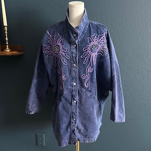 May include: A vintage denim jacket in a medium wash, featuring intricate purple floral embroidery on the front. The jacket has a high collar, button-up closure, and long sleeves. The floral design includes a central cluster of small beads.