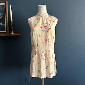 May include: Cream-colored sleeveless dress with a mandarin collar and floral embroidery. The dress features a high neckline and a knee-length hemline. The floral pattern includes red, pink, blue, and green accents. The dress is displayed on a mannequin.