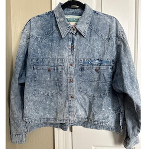 May include: A blue denim shirt with a stone wash effect. The shirt has two chest pockets, brown buttons, and the text "Code Blue" embroidered on the right pocket. The shirt has a collared neckline and long sleeves.