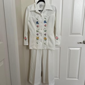 May include: A vintage white pantsuit featuring a long-sleeved button-down shirt and wide-leg pants. The shirt is adorned with colorful embroidered roses in shades of blue, pink, and yellow. The suit is made of a soft, possibly knit, fabric.