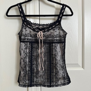 May include: Black lace camisole with a light pink lining and a matching tie. The top features delicate lace straps and trim, creating a feminine and elegant look. The camisole is hanging on a black hanger.