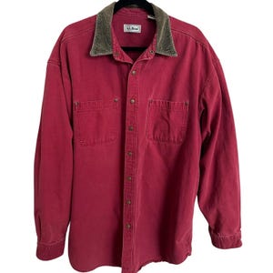 May include: A red button-down shirt with two chest pockets and a corduroy collar. The shirt has a classic design with a button-up front and long sleeves. The collar is a dark green color, and the shirt has the "L.L.Bean" brand label.