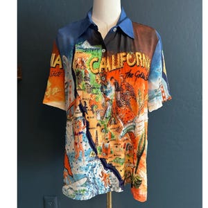 May include: A short-sleeved button-down shirt with a colorful California-themed print. The shirt features a map of California, with the words "CALIFORNIA" and "The Golden State" prominently displayed. The shirt has a blue collar and sleeves.