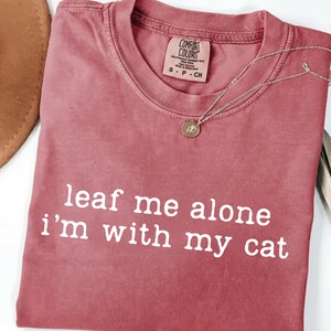 May include: A dusty rose t-shirt with the text "leaf me alone I'm with my cat" in white. A gold-colored pendant necklace and a brown hat are also visible. The shirt has a Comfort Colors tag.