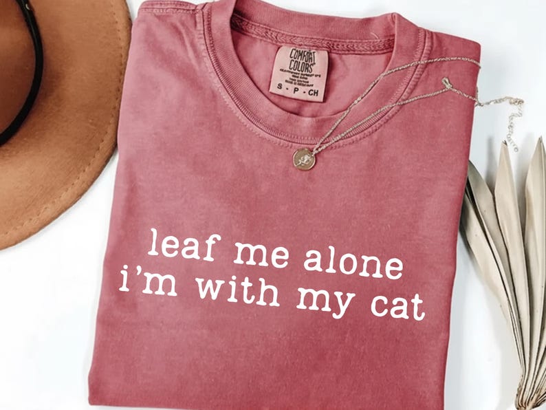 May include: A dusty rose t-shirt with the text "leaf me alone I'm with my cat" in white. A gold-colored pendant necklace and a brown hat are also visible. The shirt has a Comfort Colors tag.