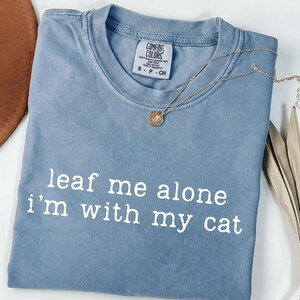 May include: A blue Comfort Colors t-shirt with the phrase "leaf me alone i'm with my cat" printed in white. A gold necklace with a pendant rests on the shirt. A brown hat and dried leaves are in the background.