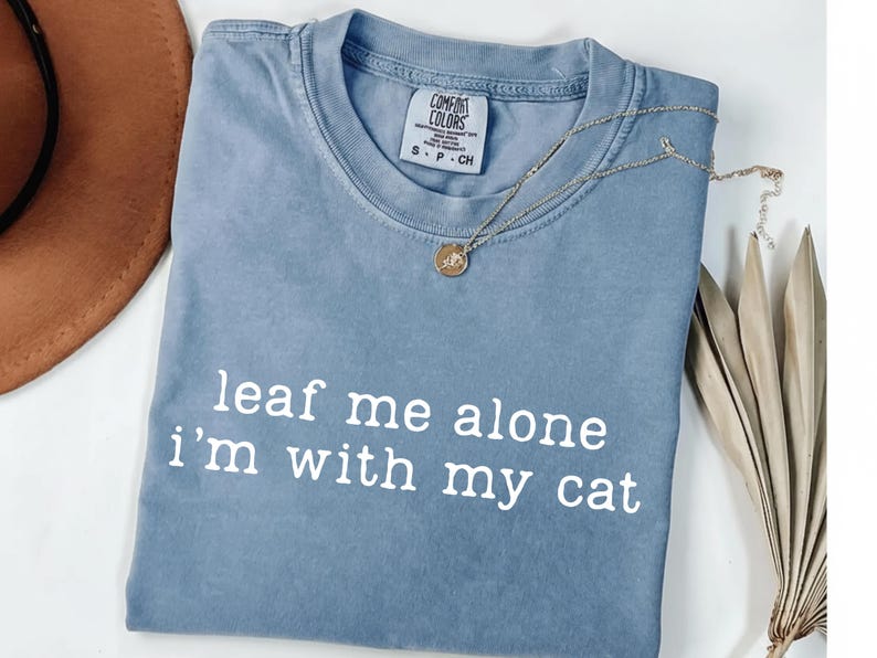 May include: A blue Comfort Colors t-shirt with the phrase "leaf me alone i'm with my cat" printed in white. A gold necklace with a pendant rests on the shirt. A brown hat and dried leaves are in the background.