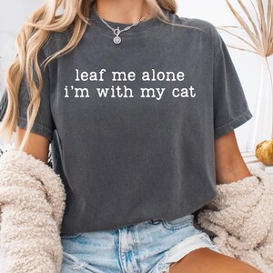 May include: Dark gray t-shirt with the text "leaf me alone i'm with my cat" in white. The shirt is worn with blue jean shorts and a cream-colored sweater. A silver necklace is visible.