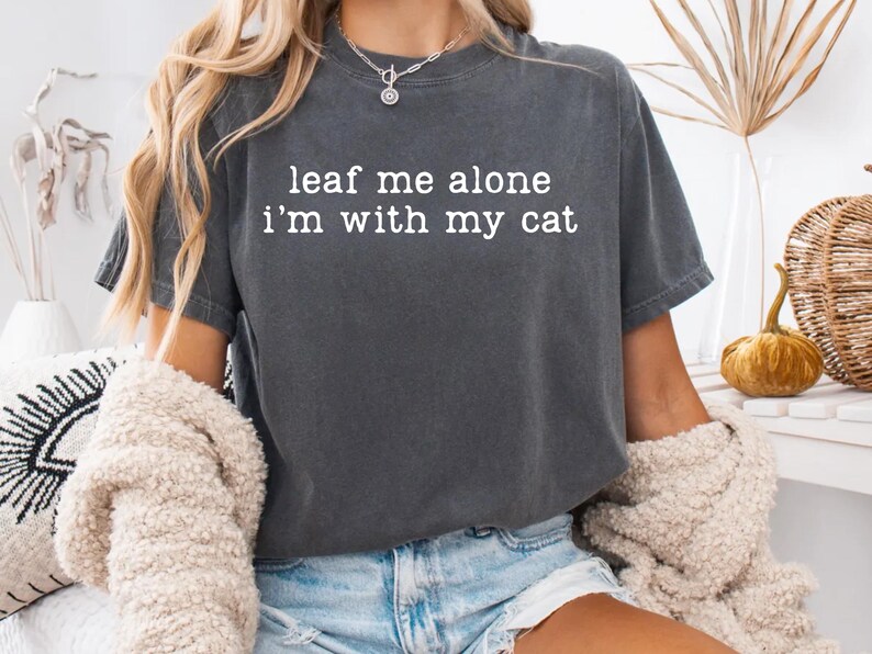 May include: Dark gray t-shirt with the text "leaf me alone i'm with my cat" in white. The shirt is worn with blue jean shorts and a cream-colored sweater. A silver necklace is visible.