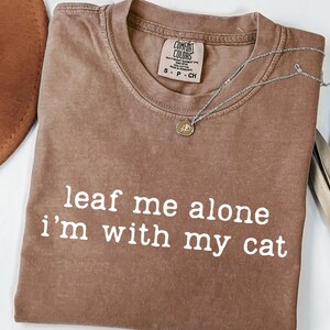 May include: A brown Comfort Colors t-shirt with the phrase "leaf me alone i'm with my cat" in white. A silver necklace with a small pendant rests on the shirt. A brown hat and dried plant are in the background.