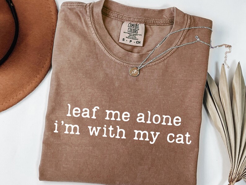 May include: A brown Comfort Colors t-shirt with the phrase "leaf me alone i'm with my cat" in white. A silver necklace with a small pendant rests on the shirt. A brown hat and dried plant are in the background.