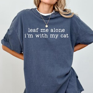 May include: A dark blue t-shirt with the text "leaf me alone I'm with my cat" in white. The shirt has a relaxed fit and is made of a soft material. The shirt is a casual wear item.