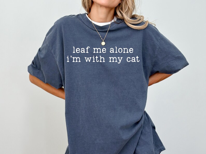 May include: A dark blue t-shirt with the text "leaf me alone I'm with my cat" in white. The shirt has a relaxed fit and is made of a soft material. The shirt is a casual wear item.