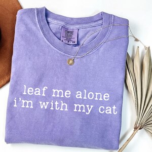 May include: A lavender Comfort Colors t-shirt with the phrase "leaf me alone i'm with my cat" in white lettering. A gold pendant necklace is draped over the shirt. A brown hat and dried palm leaves are in the background.