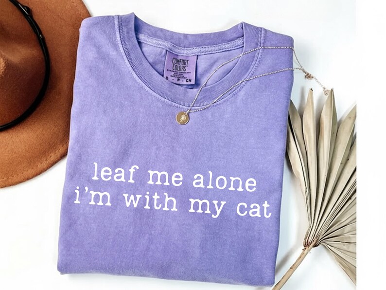 May include: A lavender Comfort Colors t-shirt with the phrase "leaf me alone i'm with my cat" in white lettering. A gold pendant necklace is draped over the shirt. A brown hat and dried palm leaves are in the background.