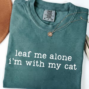 May include: A teal Comfort Colors t-shirt with the phrase "leaf me alone I'm with my cat" in white lettering. A gold necklace with a circular pendant and a brown hat are also visible.