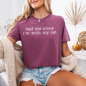May include: A maroon-colored t-shirt with the phrase "leaf me alone I'm with my cat" printed in white. The shirt is a cropped style and is being worn with denim shorts. The model is also wearing a cream-colored cardigan.