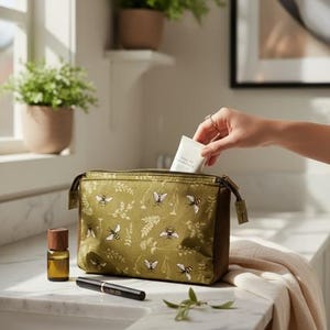 May include: An olive green cosmetic bag with a bee and floral pattern. A hand is placing a white tube into the bag. Other items on the counter include a brown essential oil bottle, a black mascara tube, and a sprig of greenery.