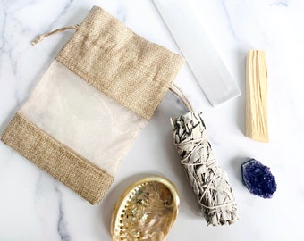 May include: A smudging kit with a white sage bundle, palo santo stick, selenite crystal, amethyst crystal, and a small burlap pouch with a clear window.