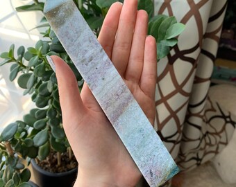 May include: A long, white, and green crystal point with a pointed tip. The crystal has a layered, striated pattern with hints of pink and brown.