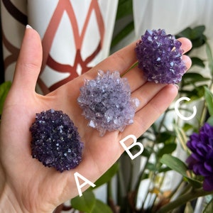 May include: Three amethyst crystal clusters, labeled A, B, and C, are displayed on a hand. The clusters are purple and have a rough, textured surface.