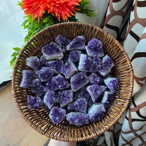 May include: A woven basket filled with raw amethyst crystals. The crystals are a deep purple color with a crystalline structure. The basket is brown and sits in front of an orange flower and green foliage.