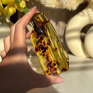 May include: A polished, translucent amber specimen featuring a golden-yellow hue with deep, dark brown organic inclusions and natural internal fractures. The stone has an irregular, elongated shape with a smooth, glossy surface texture.