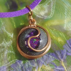 Puede incluir: Un colgante envuelto en alambre de cobre con un cabujón de resina negro y morado en su interior. El cabujón tiene un acabado brillante y un diseño yin yang.