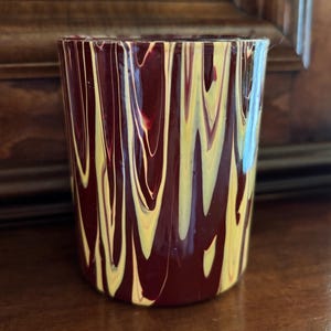May include: A cylindrical glass with a maroon base colour and a pattern of vertical yellow and white streaks. The glass is likely handmade and suitable for beverages or as a decorative item.