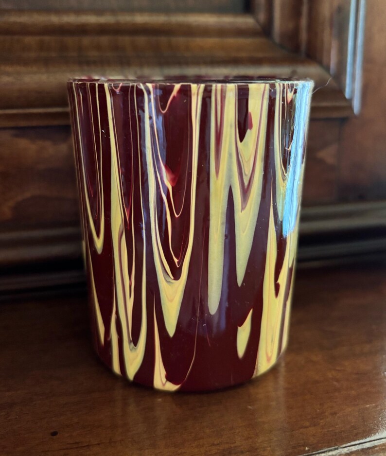 May include: A cylindrical glass with a maroon base colour and a pattern of vertical yellow and white streaks. The glass is likely handmade and suitable for beverages or as a decorative item.