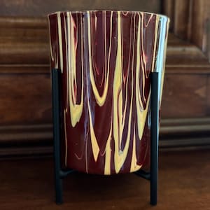 May include: A decorative glass vase with a maroon and yellow swirled pattern, set in a black metal stand. The vase has a cylindrical shape and is designed to hold flowers or other decorative items.