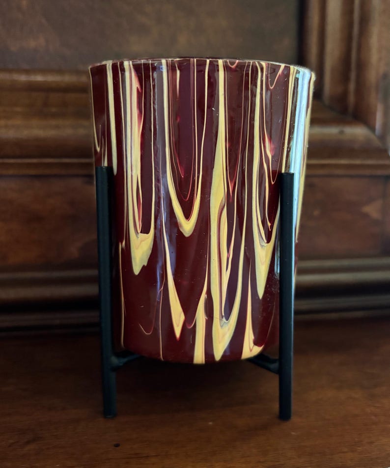 May include: A decorative glass vase with a maroon and yellow swirled pattern, set in a black metal stand. The vase has a cylindrical shape and is designed to hold flowers or other decorative items.