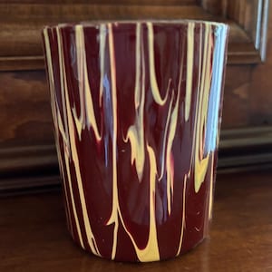 May include: A cylindrical glass with a deep burgundy base colour, adorned with vertical streaks of yellow and white. The glass has a smooth, glossy finish and is likely used for drinking beverages.