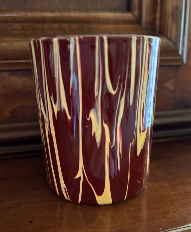 May include: A cylindrical glass with a deep burgundy base colour, adorned with vertical streaks of yellow and white. The glass has a smooth, glossy finish and is likely used for drinking beverages.