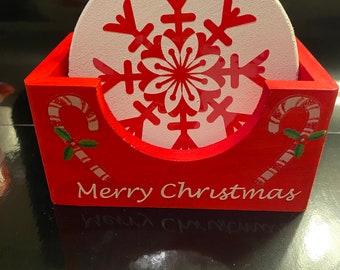 May include: Red wooden coaster holder with a white snowflake design on the coasters. The holder has the words "Merry Christmas" written on the front.